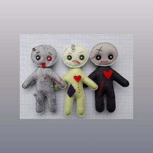 May include: Three felt voodoo dolls with stitched-on eyes, mouths, and hearts. The dolls are gray, green, and black. The gray doll has a safety pin sticking out of its head.