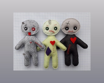 May include: Three felt voodoo dolls with stitched-on eyes, mouths, and hearts. The dolls are gray, green, and black. The gray doll has a safety pin sticking out of its head.