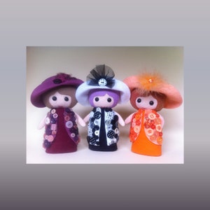 May include: Three felt dolls with button embellishments. The dolls are wearing hats and dresses. The doll on the left is wearing a purple dress with a brown hat. The doll in the middle is wearing a black and white dress with a white hat. The doll on the right is wearing an orange dress with a brown hat.