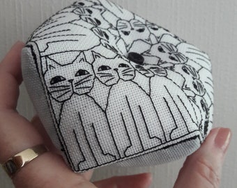 May include: A white fabric pincushion with a black outline of cats in a spiral pattern. The pincushion has a black button in the center.