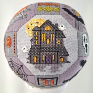 May include: A grey and black cross-stitch Halloween-themed ornament featuring a haunted house, a full moon, bats, candy corn, and the number 31.