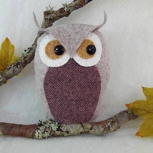 May include: A gray felt owl with brown eyes and a pink and brown herringbone pattern on its chest. The owl is perched on a branch with fall leaves.