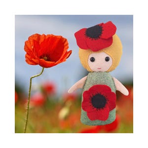 May include: A felt doll with a pale face, black bead eyes, and yellow hair adorned with a large red poppy flower. The doll wears a green felt dress featuring a prominent red poppy appliqué on the front, matching the red poppy flower nearby.