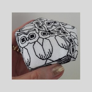 Può includere: Un cubo di tessuto bianco con gufi ricamati in nero. I gufi sono disposti in un cerchio, creando un'illusione ottica affascinante.