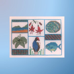 Pode incluir: Um desenho de bordado de ponto cruz que apresenta uma variedade de flora e fauna da Nova Zelândia, incluindo um pássaro azul e vermelho, uma samambaia verde, uma flor vermelha, um peixe azul e um círculo com padrão azul e verde.