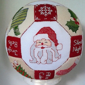 May include: A white fabric ball with a red, white and green Christmas design. The ball features a Santa Claus, a snowflake, holly, and the words "Silent Night", "Time for Bells", and "Peace on Earth".