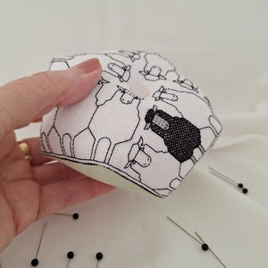 Pode incluir: Uma almofada hexagonal para alfinetes com um padrão de ovelhas. O tecido é branco com contornos pretos de ovelhas, incluindo uma ovelha preta. Um botão rosa está no topo. Vários alfinetes com cabeças pretas estão espalhados.
