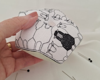 May include: A hexagonal pin cushion with a sheep pattern. The fabric is white with black outlines of sheep, and one black sheep. A pink button is on top. Several pins with black heads are scattered around.