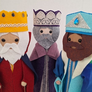 Pode incluir: Três figuras de feltro dos Três Reis Magos, cada uma com uma coroa e uma túnica únicas. As figuras têm pele castanha, barbas e estão vestidas com túnicas vermelhas, roxas e azuis. As coroas são amarelas, roxas e azuis com detalhes decorativos.