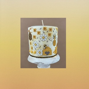 May include: A white candle with a yellow, brown, and gray bee and flower design. The candle is on a white pedestal.
