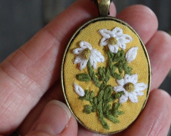May include: A yellow oval pendant necklace with a hand-embroidered design of white flowers and green leaves.