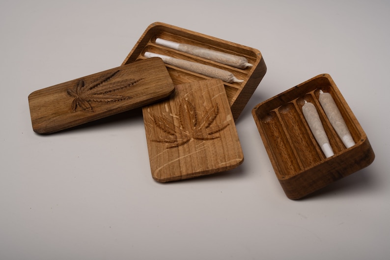 Puede incluir: Dos cajas de almacenamiento rectangulares de madera con tapas, cada una con un dise&ntilde;o de hoja de cannabis tallado en el exterior. Las cajas abiertas contienen varios conos de papel preenrollados que descansan en canales ranurados.