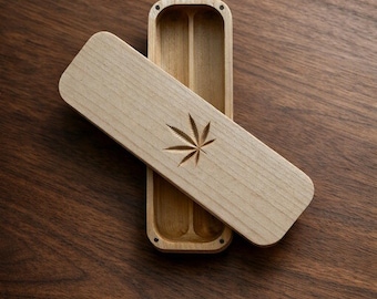 Puede incluir: Un estuche rectangular de madera clara con una tapa deslizante que presenta un diseño de hoja de cannabis grabado. El estuche está abierto, revelando dos compartimentos interiores paralelos separados por un divisor central, con pequeños imanes.
