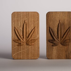 Puede incluir: Dos bloques rectangulares de madera con esquinas redondeadas, cada uno con un dise&ntilde;o de hoja de cannabis tallado. El bloque de la izquierda es de madera clara con vetas visibles, mientras que el de la derecha es de madera oscura con vetas marcadas.