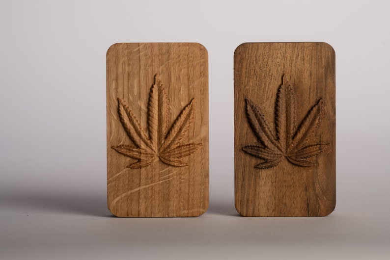 Puede incluir: Dos bloques rectangulares de madera con esquinas redondeadas, cada uno con un dise&ntilde;o de hoja de cannabis tallado. El bloque de la izquierda es de madera clara con vetas visibles, mientras que el de la derecha es de madera oscura con vetas marcadas.