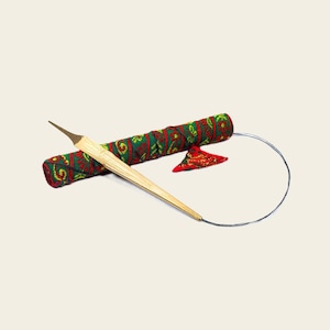 May include: A light-coloured wooden knitting needle with a pointed tip, connected to a thin grey cord. Behind the needle lies a cylindrical fabric case featuring a repeating red and green floral pattern, accompanied by a small triangular fabric tag.