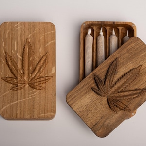 Könnte beinhalten: Zwei rechteckige Holzschachteln mit natürlicher Maserung. Jeder Deckel ist mit einem geschnitzten Cannabisblatt-Motiv versehen. Eine Schachtel ist geschlossen, während die andere offen ist und vier zylindrische Papierrollen im Inneren zeigt.