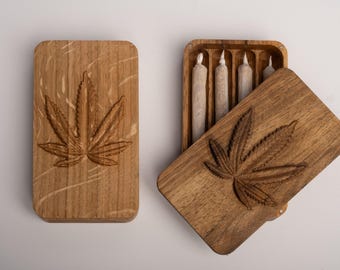Puede incluir: Dos cajas rectangulares de madera con acabado de veta natural. Cada tapa presenta un diseño tallado de hoja de cannabis. Una caja está cerrada, mientras que la otra está abierta, revelando cuatro objetos cilíndricos de papel en su interior.