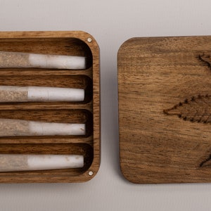 Puede incluir: Una caja rectangular de madera con cuatro ranuras internas que contienen cuatro cigarrillos liados con filtros blancos. Junto a la caja hay una tapa de madera a juego con un dise&ntilde;o de hoja de cannabis tallada.