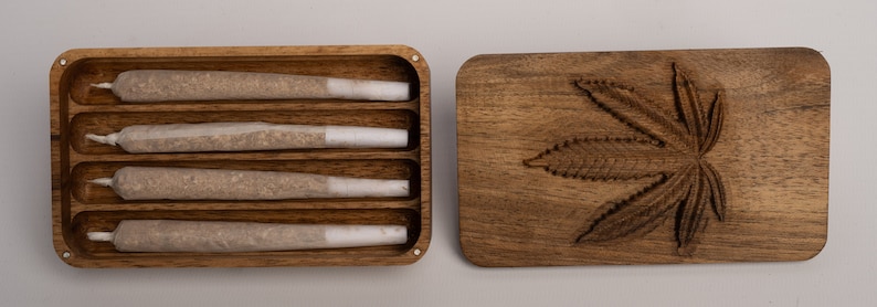 Puede incluir: Una caja rectangular de madera con cuatro ranuras internas que contienen cuatro cigarrillos liados con filtros blancos. Junto a la caja hay una tapa de madera a juego con un dise&ntilde;o de hoja de cannabis tallada.