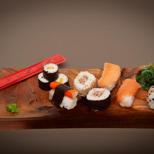 Puede incluir: Una tabla de madera con una variedad de rollos de sushi, incluyendo maki y nigiri, y una porción de ensalada de algas. Palillos chinos rojos descansan sobre la tabla. También hay una pequeña porción de wasabi.