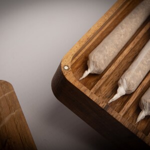 Puede incluir: Una caja de almacenamiento rectangular de madera con cierre magn&eacute;tico, que presenta m&uacute;ltiples canales interiores paralelos. Dentro de la caja hay varios objetos cil&iacute;ndricos enrollados a mano, envueltos en papel blanco fino y transl&uacute;cido con extremos retorcidos.
