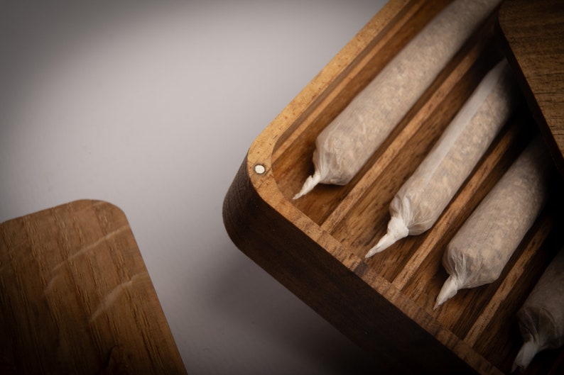 Puede incluir: Una caja de almacenamiento rectangular de madera con cierre magn&eacute;tico, que presenta m&uacute;ltiples canales interiores paralelos. Dentro de la caja hay varios objetos cil&iacute;ndricos enrollados a mano, envueltos en papel blanco fino y transl&uacute;cido con extremos retorcidos.