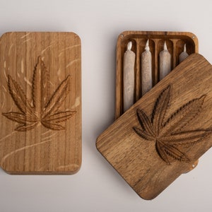 Puede incluir: Una caja de almacenamiento rectangular de madera con un dise&ntilde;o de hoja de cannabis tallado en la tapa. Una caja se muestra cerrada, mientras que la otra est&aacute; abierta, revelando cuatro objetos cil&iacute;ndricos envueltos en papel en ranuras.