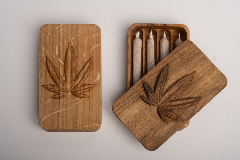 Puede incluir: Una caja de almacenamiento rectangular de madera con un dise&ntilde;o de hoja de cannabis tallado en la tapa. Una caja se muestra cerrada, mientras que la otra est&aacute; abierta, revelando cuatro objetos cil&iacute;ndricos envueltos en papel en ranuras.