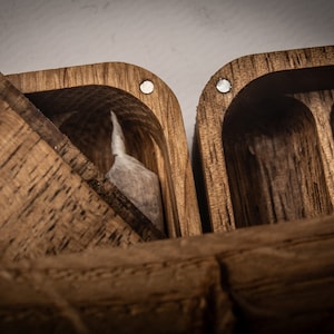 Puede incluir: Una caja de madera de color marr&oacute;n oscuro con tapa abatible y textura de grano visible. El interior presenta compartimentos tallados, uno de los cuales contiene una peque&ntilde;a bolsa rectangular blanca. Imanes circulares plateados incrustados.