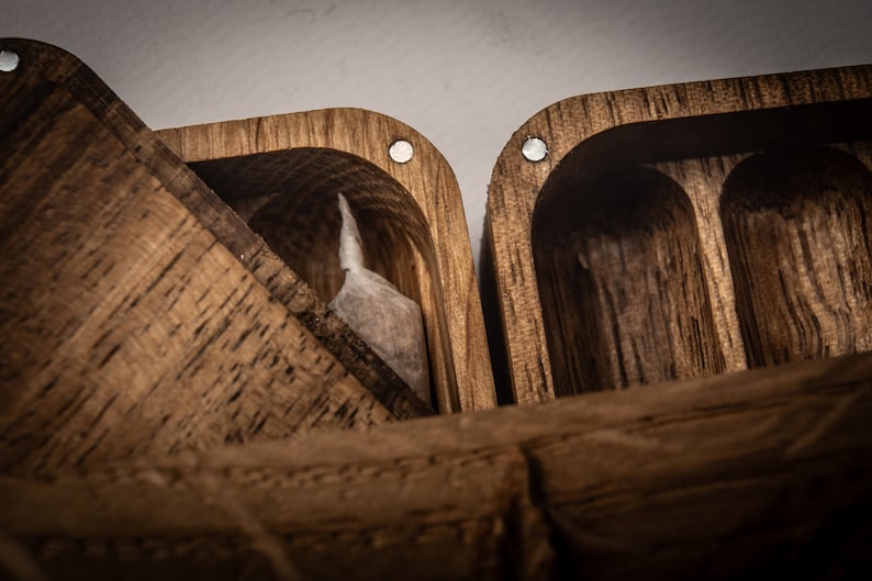 Puede incluir: Una caja de madera de color marr&oacute;n oscuro con tapa abatible y textura de grano visible. El interior presenta compartimentos tallados, uno de los cuales contiene una peque&ntilde;a bolsa rectangular blanca. Imanes circulares plateados incrustados.