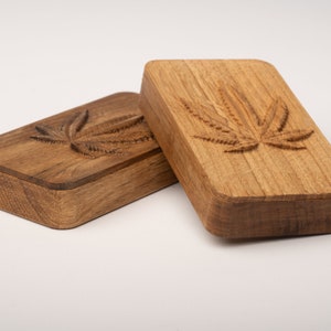 Puede incluir: Dos cajas rectangulares de madera con esquinas redondeadas, cada una con un dise&ntilde;o de hoja de cannabis tallado en la tapa. La caja de la izquierda es de madera marr&oacute;n oscura, mientras que la de la derecha tiene un acabado de madera clara natural.