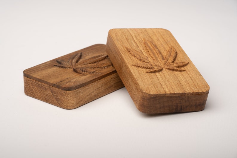 Puede incluir: Dos cajas rectangulares de madera con esquinas redondeadas, cada una con un dise&ntilde;o de hoja de cannabis tallado en la tapa. La caja de la izquierda es de madera marr&oacute;n oscura, mientras que la de la derecha tiene un acabado de madera clara natural.