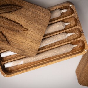 Puede incluir: Una caja de almacenamiento rectangular de madera con tapa deslizante que presenta un dise&ntilde;o de hoja de cannabis tallada. El interior contiene cuatro compartimentos ranurados, cada uno con un cilindro de papel preenrollado con filtro blanco.