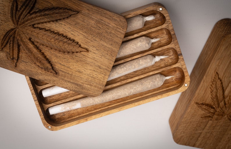 Puede incluir: Una caja de almacenamiento rectangular de madera con tapa deslizante que presenta un dise&ntilde;o de hoja de cannabis tallada. El interior contiene cuatro compartimentos ranurados, cada uno con un cilindro de papel preenrollado con filtro blanco.