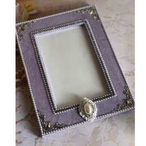 Op de afbeelding: Een lavendelkleurig fotolijstje met een parelrand en een decoratief zilveren en wit sieraad. Het lijstje heeft een witte passe-partout en is klaar om een foto te tonen.