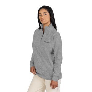 Puede incluir: Una sudadera gris con cremallera de cuarto de cuello con el texto "Keley Mom" bordado en el pecho. La sudadera tiene un cuello alto y una cremallera plateada. La modelo lleva pantalones beiges.