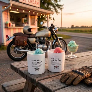 May include: Two white candles with ice cream-themed scents, topped with scoops of blue and pink ice cream, sit on a wooden picnic table. The candles are labeled "Ride to Ice Cream." A vintage motorcycle and ice cream stand are in the background.