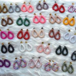 Könnte beinhalten: Eine Sammlung von tropfenförmigen Ohrringen mit Perlen in verschiedenen Farben, darunter Blau, Gold, Rosa, Rot und Grün. Jedes Paar hat einen goldfarbenen Stecker. Die Ohrringe werden auf einer weißen, flauschigen Oberfläche präsentiert.