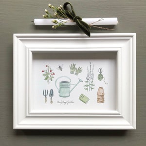 May include: A white framed print with a watercolor illustration of gardening tools and plants. The print says "The Cottage Garden" at the bottom.