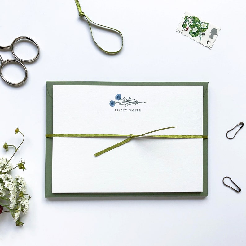 May include: A white card with a green border and a green ribbon tied around it. The card has a blue floral design and the text "Poppy Smith" printed on it.