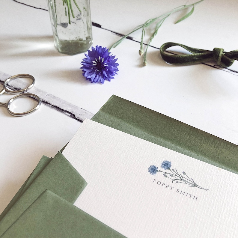 May include: A white card with a green envelope. The card has a small illustration of blue flowers and the text "Poppy Smith".