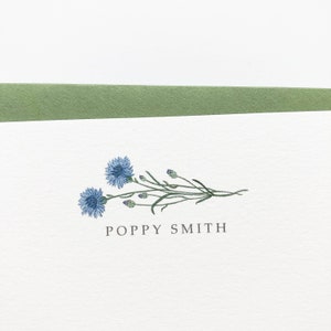 May include: A white card with a green border at the top. The card features a blue flower illustration with green stems and leaves. The text "POPPY SMITH" is printed in black below the illustration.