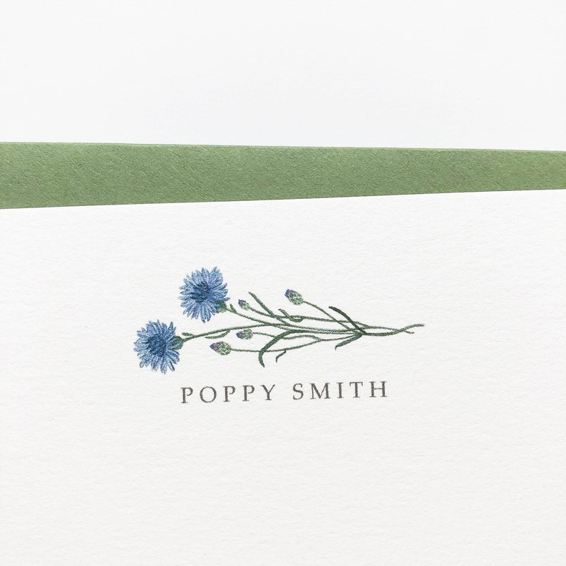 May include: A white card with a green border at the top. The card features a blue flower illustration with green stems and leaves. The text "POPPY SMITH" is printed in black below the illustration.