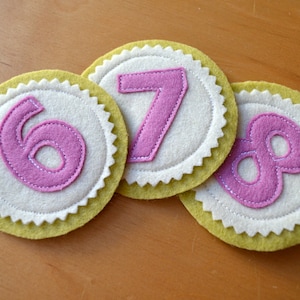 Könnte beinhalten: Drei Filz-Kreise mit den pinkfarbenen Zahlen 6, 7 und 8 auf weißem Hintergrund. Die Kreise sind gelb mit einem gewellten Rand.