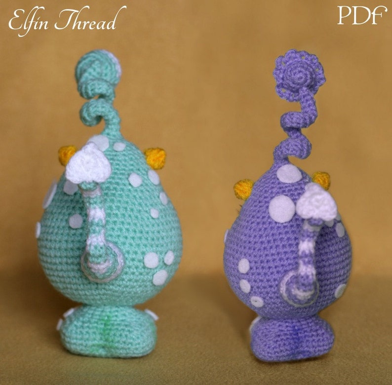 May include: Two crocheted amigurumi toys, one green and one purple, with white spots and yellow accents. Both toys have a curled tail and a white cloud-like head.