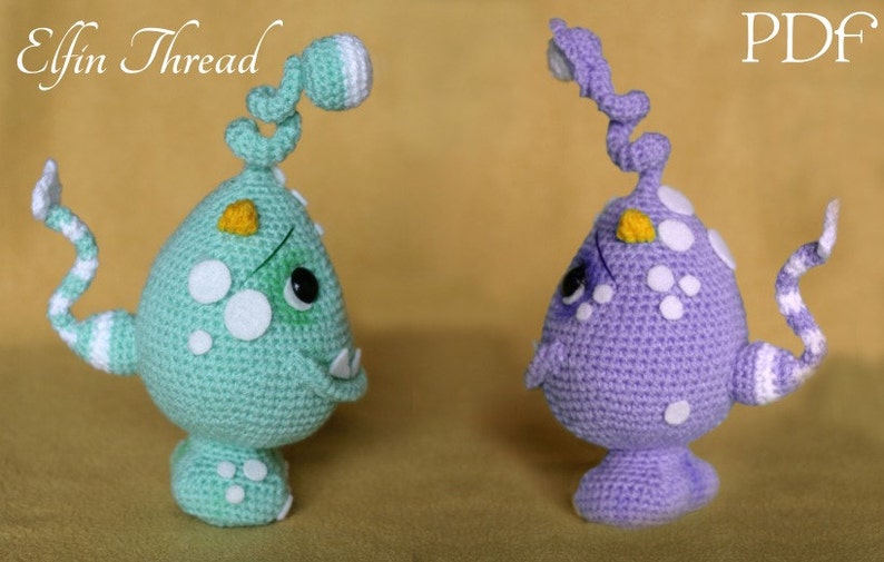May include: Two crocheted amigurumi monsters, one green and one purple, with white spots and yellow accents. The monsters have long, curled tails and are standing on a beige surface. The text "Elfin Thread" is visible in the top left corner of the image, and "PDF" is visible in the top right corner.