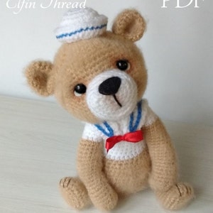 May include: A crocheted teddy bear wearing a white sailor hat with blue stripes and a red bow tie. The bear is sitting on a light wood surface.