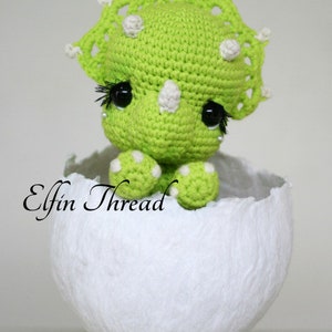 May include: A green crocheted dinosaur with white accents and large black eyes sits inside a white crocheted egg. The dinosaur has a frill around its head and a small horn on its nose. The text "Elfin Thread" is visible in the image.