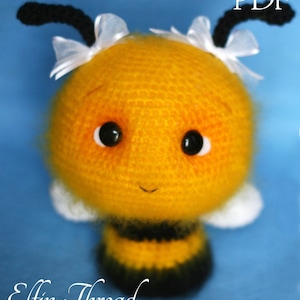 May include: A crocheted bee toy with a yellow body, black stripes, and white wings. The bee has black antennae, white bows, and black eyes. The bee is smiling and has a fuzzy texture.